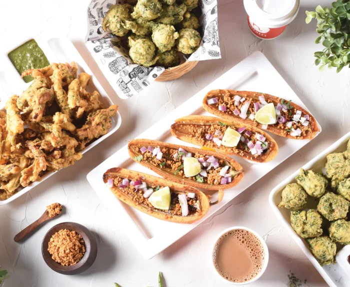 An assortment of Indian street food from Neehee's displayed on a white table. The dishes include crispy pakoras with green and tamarind chutneys, a basket of spinach fritters, four stuffed dabeli buns garnished with onions and lime wedges, and another tray of green fritters with dipping sauces. A cup of chai and a takeaway coffee cup are also visible, along with fresh coriander and a small bowl of dry chutney.