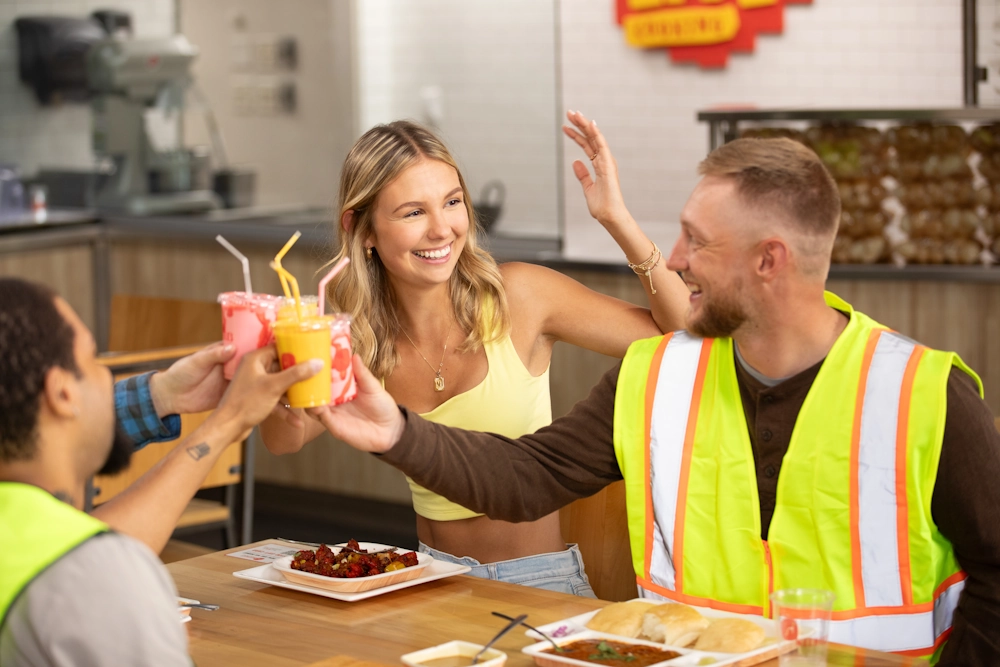 A group of happy customers toasting with colorful drinks like lassi while enjoying a variety of Indian street food dishes such as Pav Bhaji and Manchurian at a Neehee's restaurant.