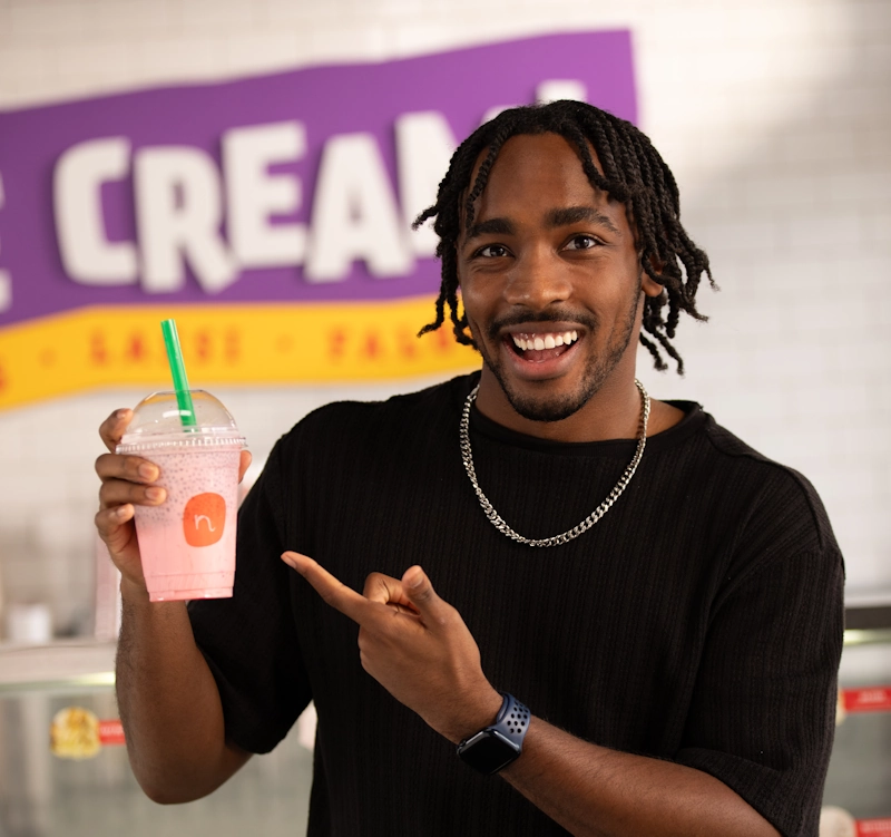 A happy customer at a Neehee's restaurant smiling and pointing at his Falooda or milkshake. The pink-colored drink is in a clear cup with the Neehee's logo.