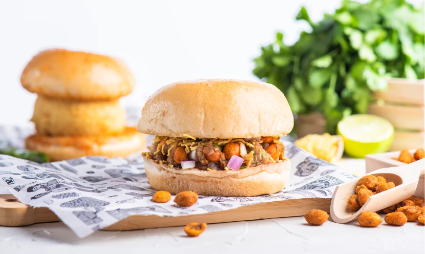 Close-up of Neehee's Dabeli stuffed pav, garnished with red onions, masala peanuts, and fine sev, on patterned wrapping paper.