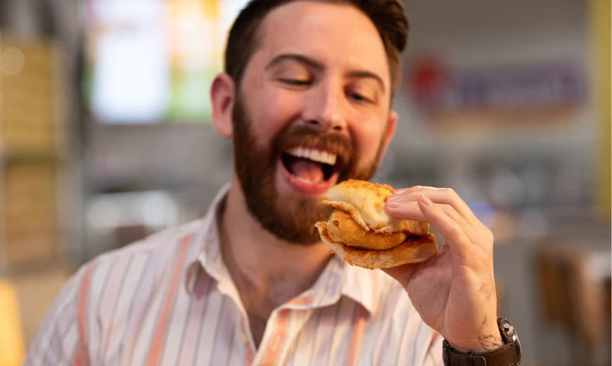 Bearded man smiling while enjoying a bite of Neehee's Vada Pav (spicy potato fritter sandwich) in the restaurant.