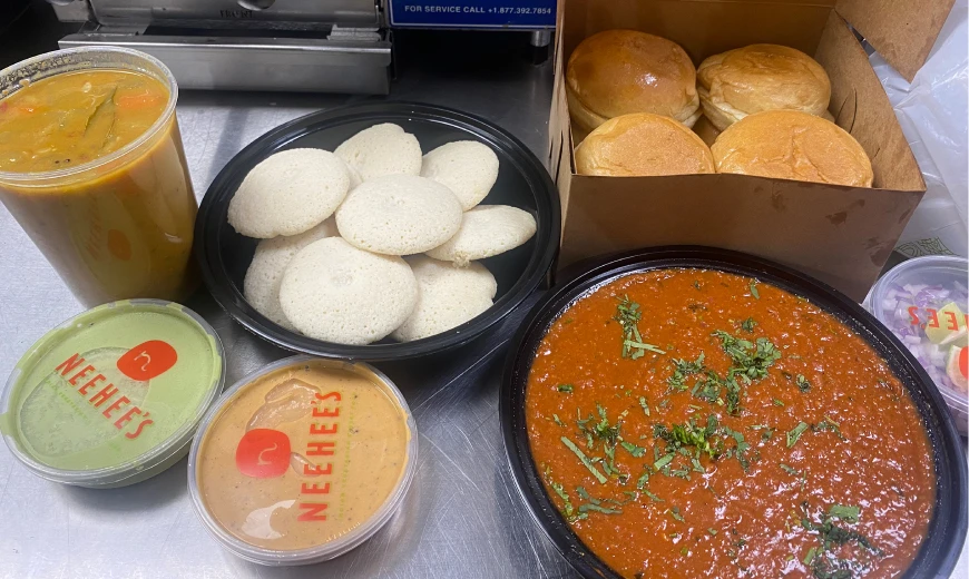 Indian meal combo with soft idlis, spicy sambar soup, tangy chutneys, buttery pav buns, and rich red curry, served in a takeout box from Neehee's on a metal surface.