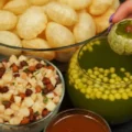 A hand holding a green pani puri filled with chickpeas and tamarind water, next to bowls of crispy puris, spiced potato-chickpea filling, tangy green pani, and red chutney on a black surface.