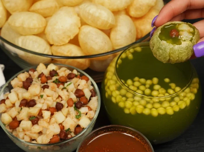 A hand holding a green pani puri filled with chickpeas and tamarind water, next to bowls of crispy puris, spiced potato-chickpea filling, tangy green pani, and red chutney on a black surface.