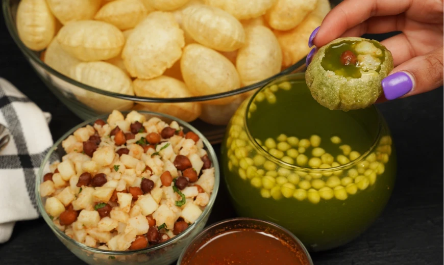 A hand holding a green pani puri filled with chickpeas and tamarind water, next to bowls of crispy puris, spiced potato-chickpea filling, tangy green pani, and red chutney on a black surface.